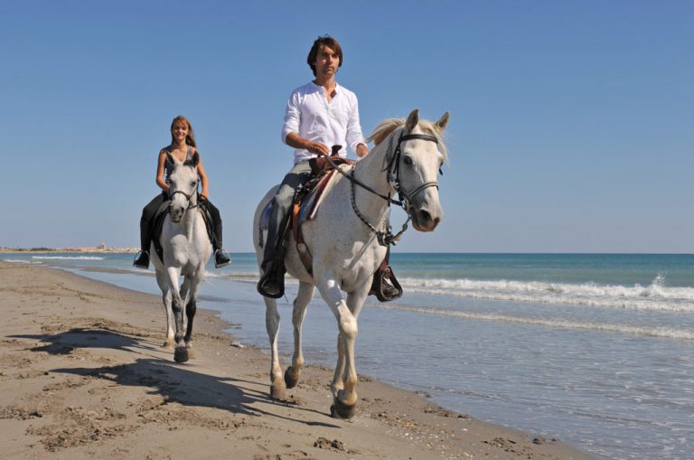 Un chico y una chica subidos a dos caballos blancos, paseando en una playa.